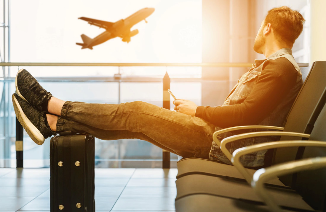 Man sitting on airport seat with legs stretched out on top of black suitcase while he is watching airplanes depart through airport window. 